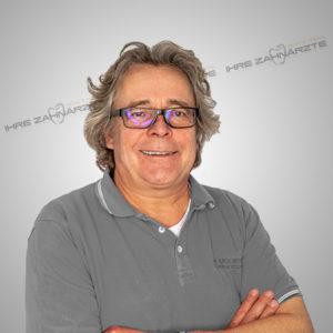 Lächelnder Zahnarzt in grauem Polohemd mit Brille, präsentiert sich vor einem neutralen Hintergrund. Ideal für die Website einer Zahnarztpraxis.