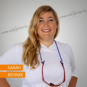 Portrait von Sarah Behnke, einer Zahnärztin, die lächelnd in einem weißen Oberteil posiert. Sie trägt eine Brille um den Hals und hat lange, blonde Haare. Der Hintergrund ist neutral gehalten, um den Fokus auf die Zahnärztin zu lenken.
