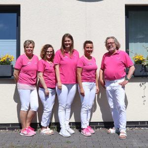 Teamfoto einer freundlichen Gruppe von fünf Personen in pinken Polohemden und weißen Hosen vor einem Gebäude, das eine einladende Atmosphäre ausstrahlt. Ideal für die Präsentation des Unternehmens und der Mitarbeiter.