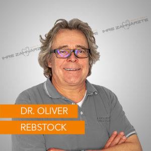 Dr. Oliver Rebstock, Zahnarzt mit lächelndem Gesicht und grauen Haaren, trägt eine graue Polo-Shirt und eine Brille. Im Hintergrund ist das Logo "Ihre Zahnärzte" zu sehen.