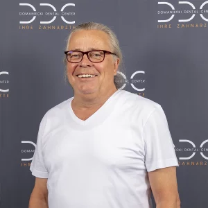 Lächelnder Zahnarzt in einem weißen T-Shirt vor einem Hintergrund des Domaniecki Dental Centers mit dem Slogan Ihre Zahnärzte.