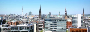 Panoramaaufnahme der Hamburger Skyline mit modernen Gebäuden und historischen Kirchtürmen, darunter der Michel und der Fernsehturm, bei klarem Himmel.