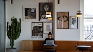 Frau sitzt an einem Tisch in einem modernen Café und arbeitet an einem Laptop. An der Wand hängen verschiedene Kunstwerke, darunter Bilder von Picasso und Tapies. Ein großer Kaktus steht neben ihr. Die Atmosphäre ist hell und einladend.